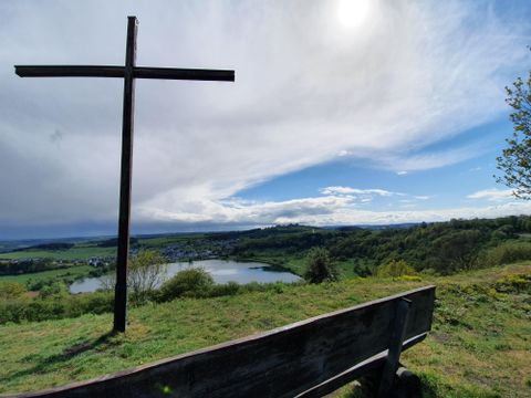 Ein Holzsteg führt über einen ruhigen See, umgeben von grünem Gras und Bäumen. Der Himmel ist klar mit einigen Wolken.