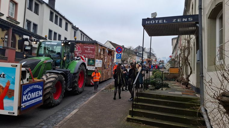 Eine Straße mit einem Traktor und einem festlich dekorierten Wagen. Menschen stehen auf dem Gehweg in der Nähe des Hotels ZENS.