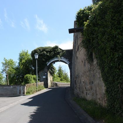 Eine malerische Straße führt durch ein historisches Tor. Umgeben von üppigem Grün und klarblauem Himmel, strahlt die Szene Ruhe und Schönheit aus.