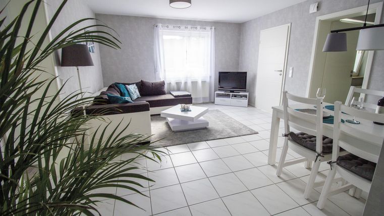 Ein modernes Wohnzimmer mit hellen Wänden und einem großen Fenster. Es gibt eine gemütliche Couch, einen Fernseher und einen Esstisch mit Stühlen.