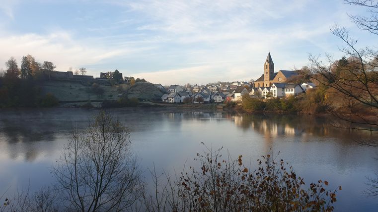 Eine ruhige Landschaft mit einem See und einem Blick auf ein malerisches Dorf. Im Hintergrund ist ein Kirchturm und eine bewaldete Hügelkette zu sehen.