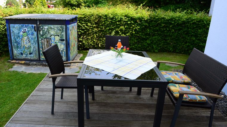 Ein gemütlicher Gartentisch mit Stühlen und einer Tischdecke. Im Hintergrund ist eine grüne Hecke und ein bunt bemalter Schuppen zu sehen.