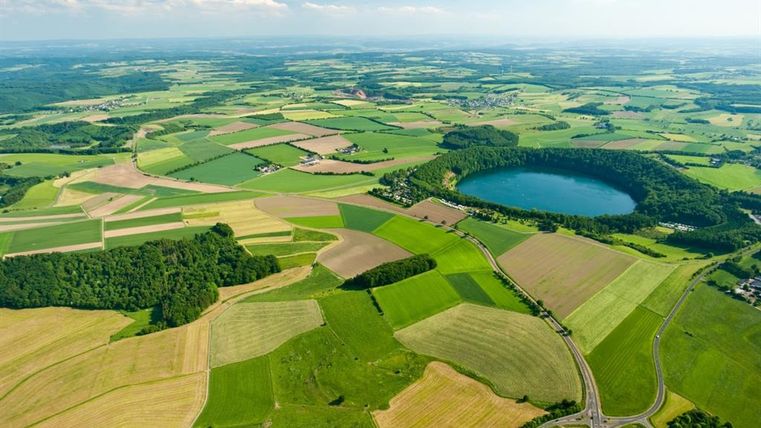 Eine weite Landschaft mit grünen Feldern und einem blauen See in der Mitte. Sanfte Hügel und ein klarer Himmel vervollständigen die malerische Aussicht.