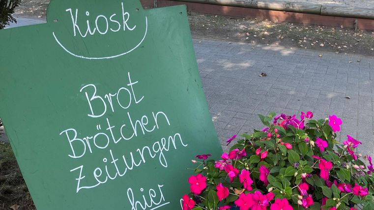 Ein Kiosk-Schild mit der Aufschrift „Brot, Brötchen, Zeitungen“. Daneben blühen pinkfarbene Blumen.