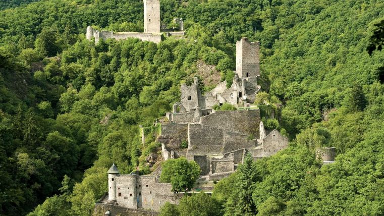 Eine malerische Burgruine, umgeben von grünen Wäldern. Im Hintergrund erheben sich weitere Ruinen auf dem Hügel.