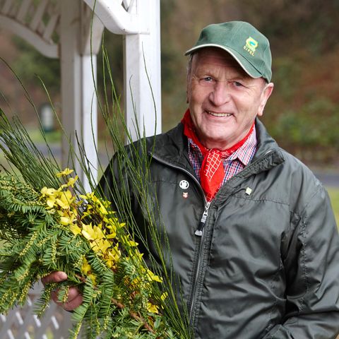 Ein älterer Mann trägt eine grüne Jacke und einen grünen Hut. Er hält einen Strauß mit gelben Blumen und grünen Zweigen in der Hand.