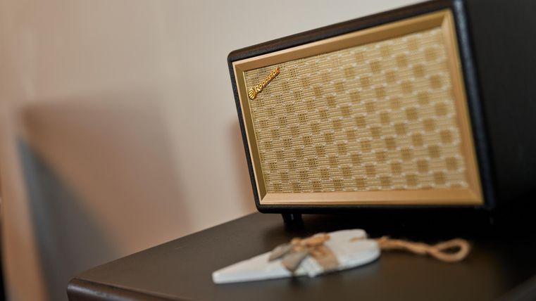 Ein eleganter Lautsprecher mit einem goldenen Gitter und schwarzem Gehäuse. Daneben liegt ein kleiner Anhänger auf einer dunklen Oberfläche.