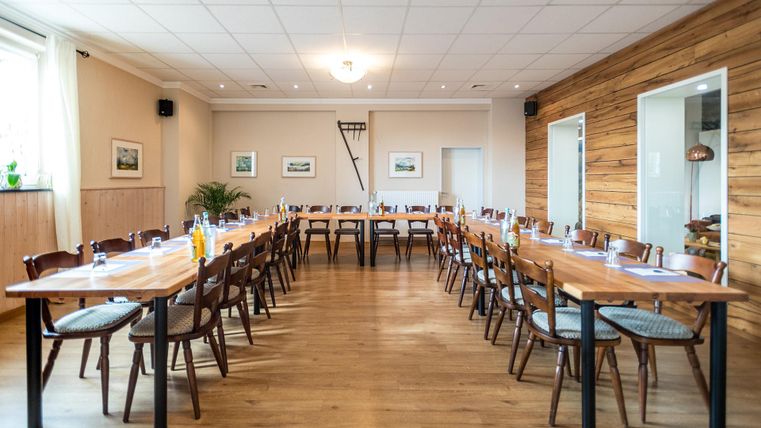 Ein helles Besprechungszimmer mit langen Tischen und vielen Stühlen. Die Wände sind teilweise aus Holz und es gibt Pflanzen zur Dekoration.