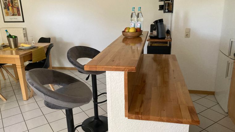 Eine moderne Küche mit einer Holzbar und hohen Stühlen. Darauf stehen Flaschen Wasser und ein Obstkorb.