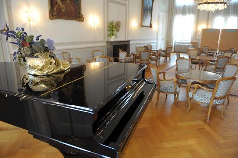 Ein elegantes Klavier steht in einem stilvollen Raum mit Holzboden. Rund um das Klavier befinden sich gemütliche Tische und Stühle.