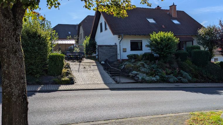 Ein gemütliches Einfamilienhaus mit einem gepflegten Garten. Die Straße ist ruhig und von Bäumen gesäumt.