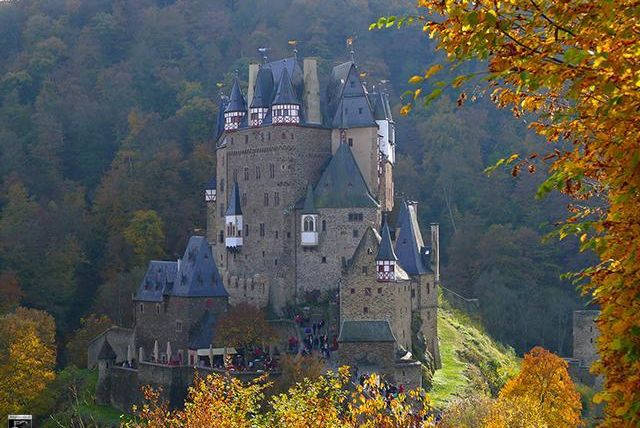 Eine imposante Burg umgeben von bunten Herbstbäumen. Die Landschaft zeigt eine malerische Kulisse mit sanften Hügeln.