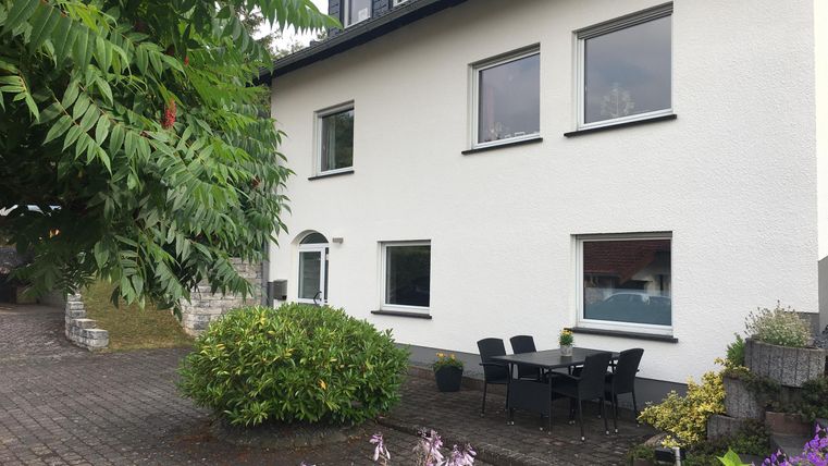 Ein modernes Haus mit weißen Wänden und großen Fenstern. Vor der Haustür befindet sich ein kleiner Garten mit einer Sitzgruppe.