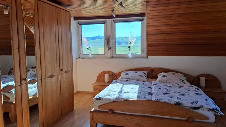 Ein gemütliches Schlafzimmer mit einem Holzbett und einer weißen Bettwäsche. Zwei Fenster bieten einen Blick auf eine grüne Landschaft.