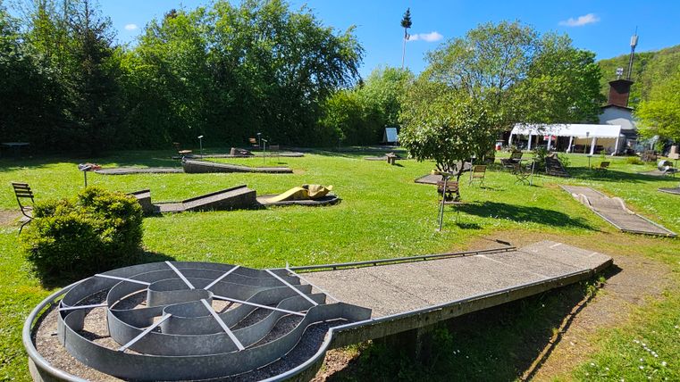 Eine Minigolfanlage mit mehreren Bahnen und viel grünem Rasen. Im Hintergrund sind Bäume und eine überdachte Sitzgelegenheit zu sehen.