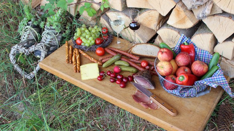 Ein rustikaler Tisch mit einer Auswahl an frischem Obst, Gemüse, Käse und Wurst. Im Hintergrund sind Holzscheite und ein Glas Wein zu sehen.