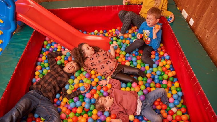 Eine Gruppe von Kindern spielt in einem farbenfrohen Bällebad. Im Hintergrund ist eine Rutsche zu sehen.