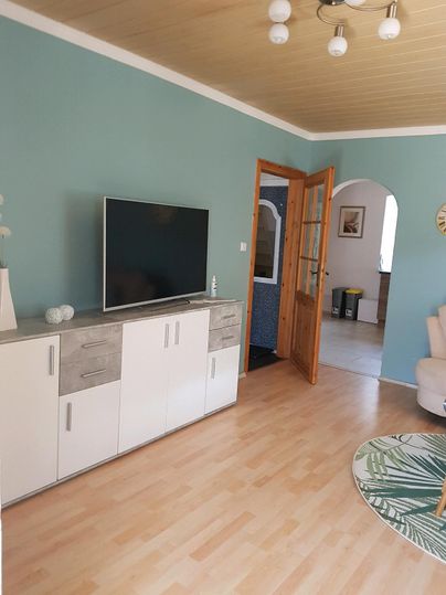Ein helles Wohnzimmer mit einer blauen Wand und einer modernen Fernseherwand. Im Hintergrund ist eine offene Tür, die zu einem angrenzenden Raum führt.