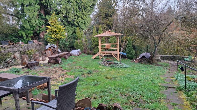 Ein Garten mit grüner Wiese und mehreren Bäumen. Im Hintergrund steht eine Holzpergola, und ein Tisch mit Stühlen ist im Vordergrund sichtbar.