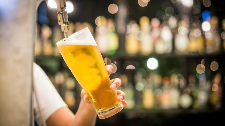 Ein Glas frisch gezapftes Bier wird serviert. Im Hintergrund sind unscharfe Flaschen zu sehen.