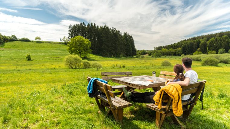 Ein Paar sitzt auf einer Bank an einem Holztisch in einer grünen Landschaft mit Bäumen im Hintergrund.