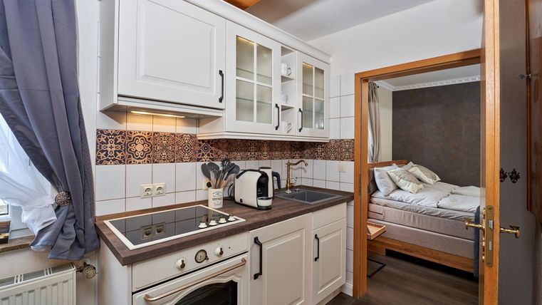 Eine helle Küche mit weißen Schränken und modernen Geräten. Durch eine Tür ist ein Schlafzimmer sichtbar.