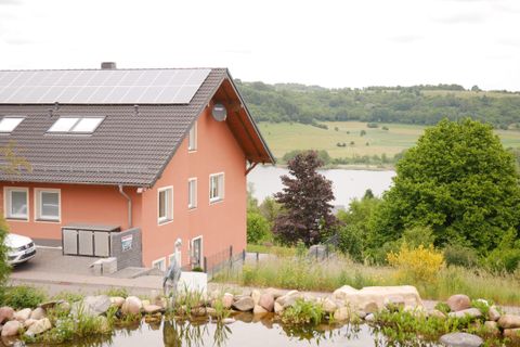 Das Bild zeigt ein modernes Haus mit einer Solaranlage auf dem Dach. Im Hintergrund erstreckt sich eine grüne Landschaft mit einem See.