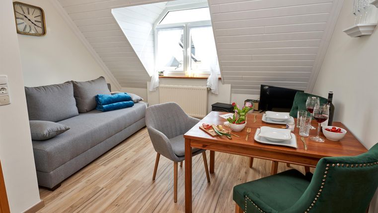 Ein gemütlicher Wohnbereich mit einer grauen Couch und einem kleinen Esstisch. Der Raum hat große Fenster und eine angenehme, einladende Atmosphäre.