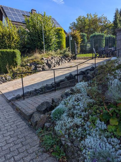 Eine gepflasterte Auffahrt führt zu einem Garten mit Sträuchern und Blumen. Im Hintergrund sind ein zaun und ein Haus mit Solarpanelen zu sehen.