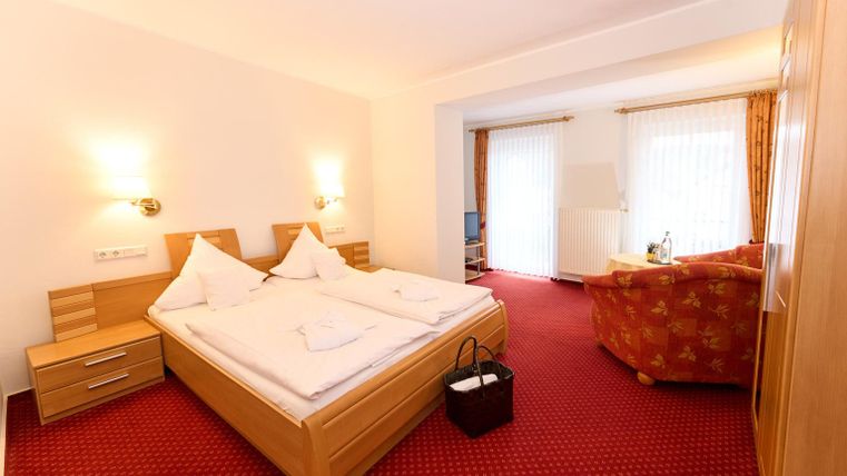 Ein gemütliches Hotelzimmer mit einem Doppelbett und einer roten Teppichfläche. Es gibt einen Wohnbereich mit einem Sofa und viel Tageslicht.