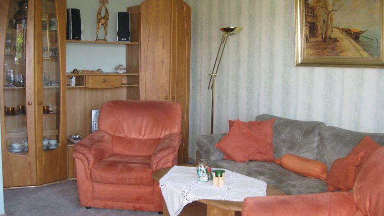 Ein gemütliches Wohnzimmer mit einer grauen Couch und zwei orangenen Sesseln. An der Wand hängt ein Gemälde, und im Raum sind Holzschränke mit Dekorationsgegenständen.