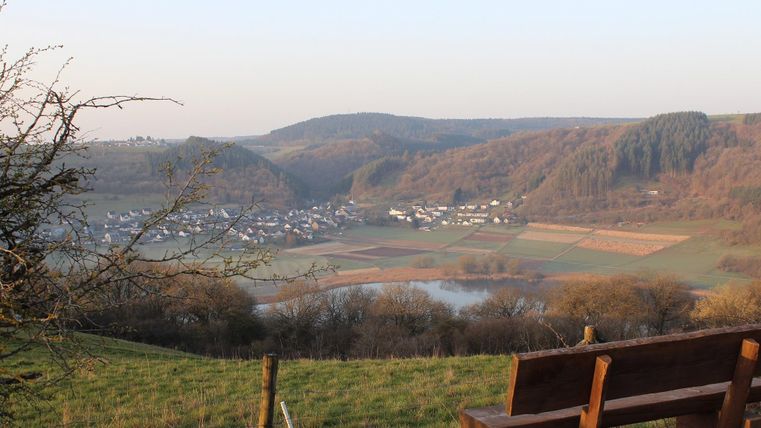 Eine malerische Landschaft mit sanften Hügeln und einem kleinen Dorf im Tal. Im Vordergrund steht eine Holzbank, die einen schönen Blick auf die Umgebung bietet.