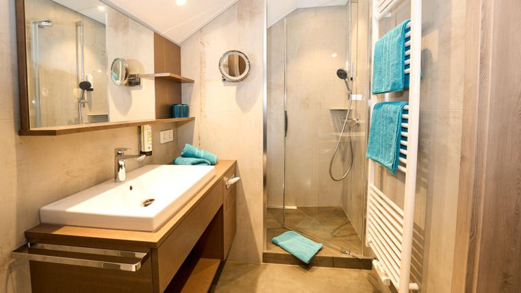 Ein modernes Badezimmer mit einer Dusche und einem Doppelwaschbecken. Die Wände sind in neutralen Tönen gehalten, ergänzt durch handtücher in Türkis.