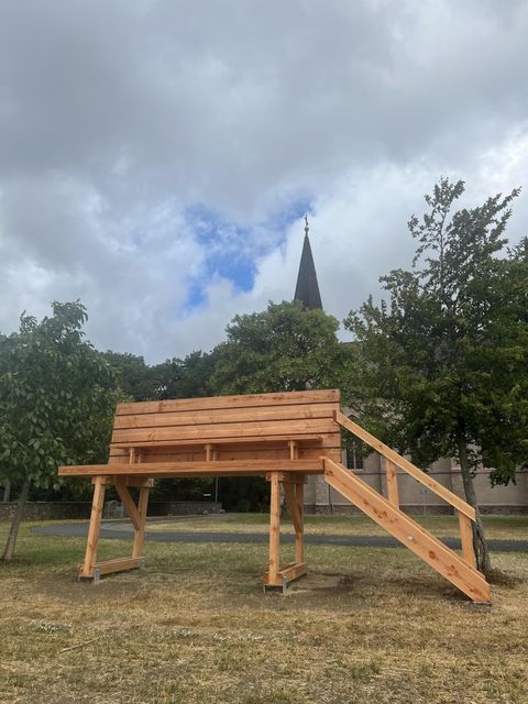 Eine hölzerne Aussichtsbank steht in einem Garten. Im Hintergrund ist ein Kirchturm und bewaldete Bereiche zu sehen.