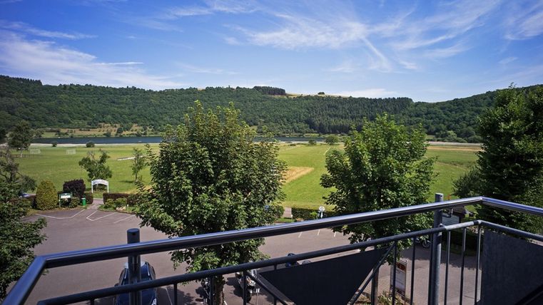 Ein schöner Blick von einem Balkon auf eine grüne Landschaft mit Bäumen. Im Hintergrund sind sanfte Hügel und ein blauer Himmel zu sehen.