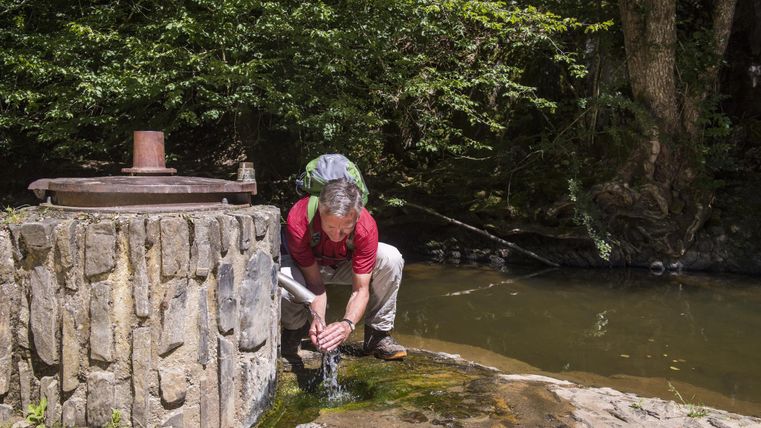Ein Mann beugt sich über einen Brunnen und schöpft Wasser. Im Hintergrund sind Bäume und ein ruhiger Fluss zu sehen.