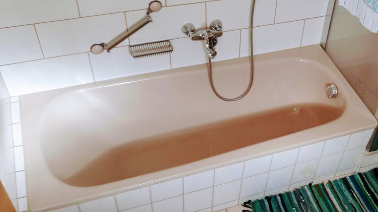 Eine einfache Badewanne auf weißen Fliesen. Daneben liegt ein gestreifter Teppich.