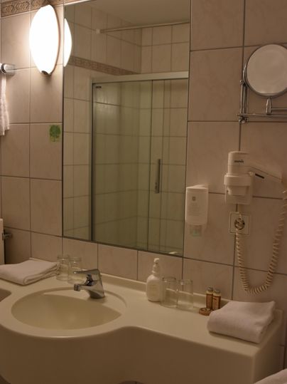 Ein modernes Badezimmer mit einem Waschbecken und einem großen Spiegel. Es gibt eine Dusche und einige Toilettenartikel auf der Ablage.