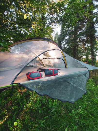 Ein Campingzelt auf einer Wiese, umgeben von Bäumen. Zwei Rucksäcke liegen auf der Zeltfläche.