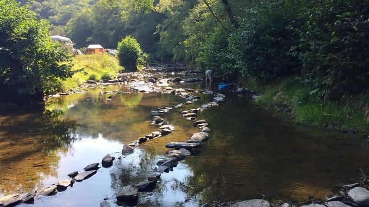 Ein ruhiger Fluss verläuft durch einen bewaldeten Bereich, mit flachen Steinen, die im Wasser liegen. Im Hintergrund sind Bäume und einige Zelte sichtbar.