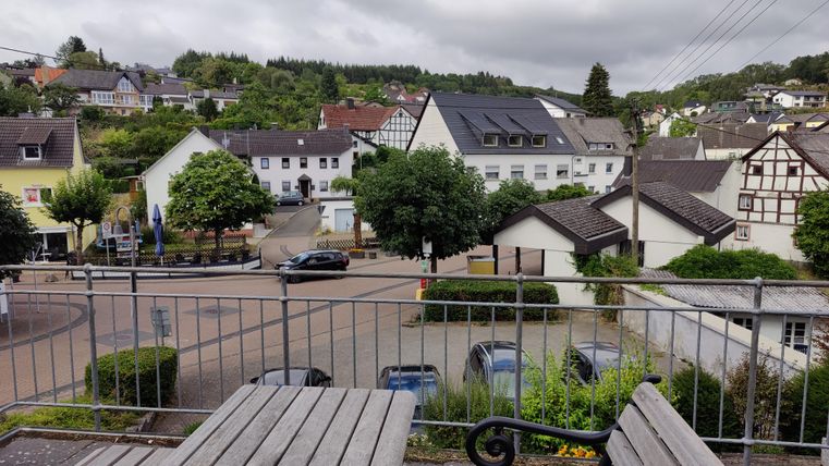 Ein ruhiger Blick auf ein kleines Dorf mit traditionellen Häusern. Die Gegend ist grün und es gibt einige Bäume sowie parkende Autos.