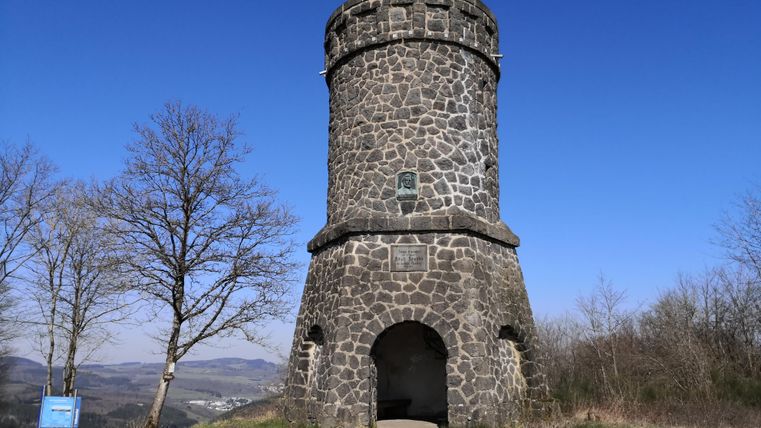 Ein steinerner Aussichtsturm steht in einer ruhigen Landschaft. Der Himmel ist klar und blau, und es gibt einige Bäume in der Nähe.