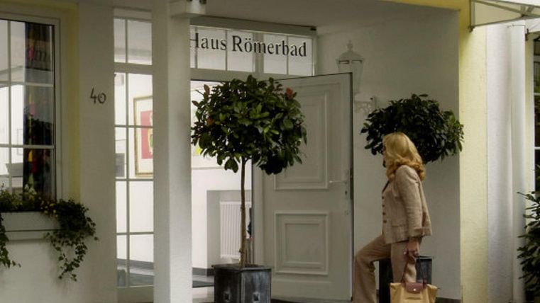Eine elegante Fassade des Hauses Römerbad mit einer Frau, die zur Tür geht. Üppige Pflanzen dekorieren den Eingangsbereich.