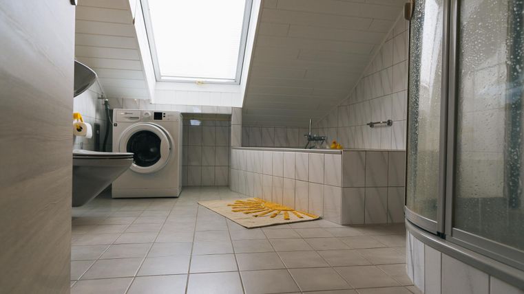 Ein modernes Badezimmer mit Dachfenster, einer Waschmaschine und einer Dusche. Der Raum ist hell und hell gefliest, mit einem gelben Teppich auf dem Boden.