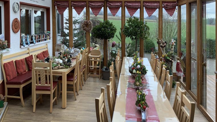 Ein helles Wintergarten mit langen Tischen und bunten Stühlen. Blumenarrangements und eine grüne Aussicht schaffen eine einladende Atmosphäre.