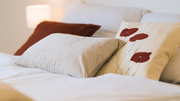 Ein gemütliches Bett mit mehreren Kissen. Ein Kissen hat ein Blumenmuster und die Bettwäsche ist hell und freundlich.