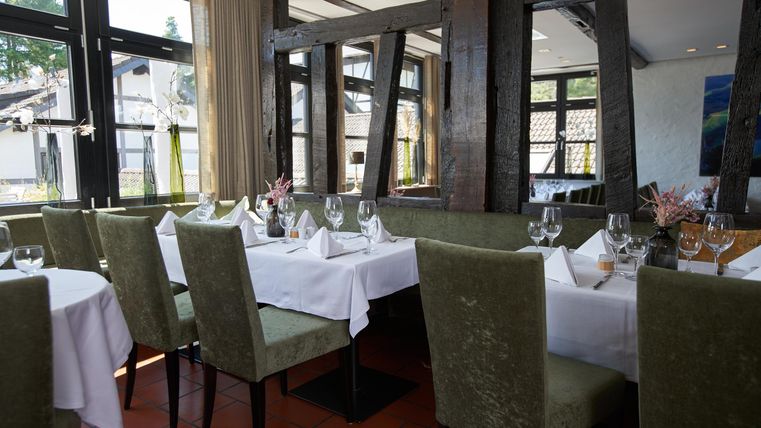 Ein elegantes Restaurant mit stilvollen Tischen und grünen Stühlen. Die großen Fenster lassen viel Licht herein und bieten einen Blick auf die Umgebung.