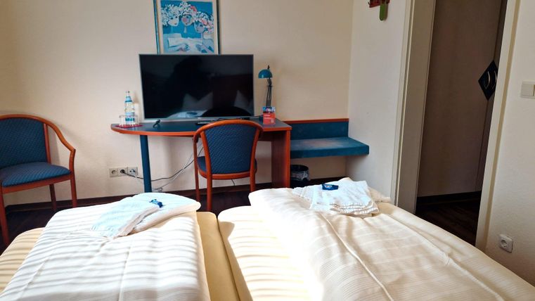 Ein einfaches Hotelzimmer mit zwei getrennten Betten. Ein Tisch mit einem Fernseher und Stühlen ist ebenfalls vorhanden.