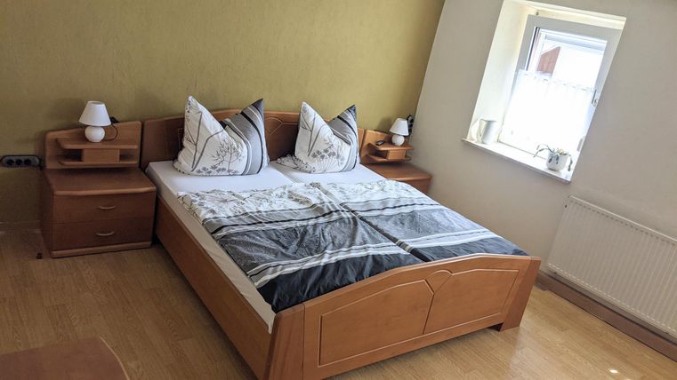 Ein gemütliches Schlafzimmer mit einem Doppelbett und zwei Nachtkästchen. Das Zimmer hat eine helle Wand und ein Fenster, das Tageslicht hereinlässt.