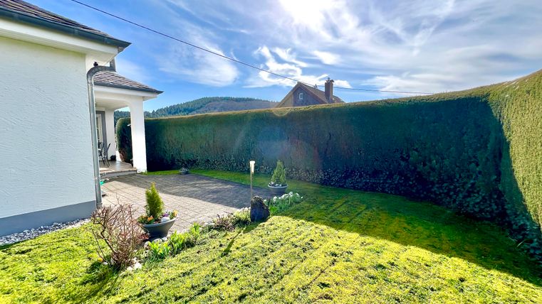 Ein gepflegter Garten mit grünem Rasen und einer hohen Hecke umgeben. Die Sonne scheint hell und der Himmel ist blau mit einigen Wolken.